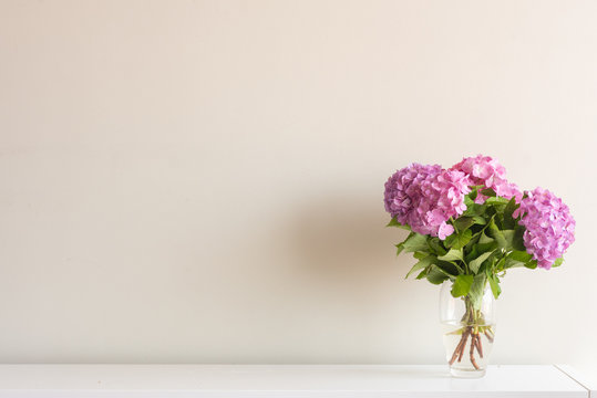 Pink hydrangea flowers with green leaves in glass vase on white side board against neutral wall background