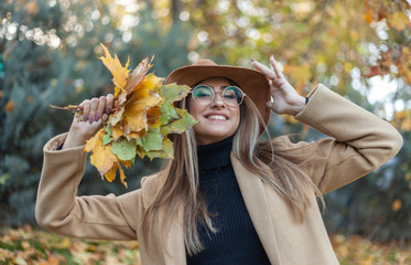 Young funny woman dressed in a coat and hat holds an armful of fallen leaves in the autumn park. Autumn fun