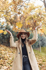 Young funny woman dressed in a coat and hat throws up fallen leaves in the autumn park. Autumn fun