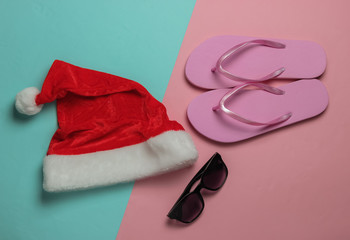 Santa hat, flip flops and sunglasses on pink blue background. New Year, Christmas holiday in warm...