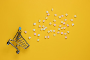 Lot of mini marshmallows and shopping trolley on yellow background. Top view