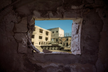 Peeping through a hideaway wall hole to see a tank preparing to shoot at a demolished stone building