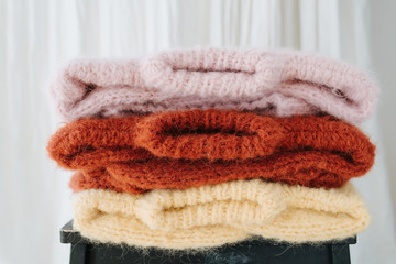 Fototapeta na wymiar Three folded multi-colored knitted sweaters lie on a stool in front of a white curtain. Laundry day. Close up.