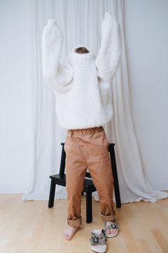 Funny little girl struggling to put on white fluffy knitted sweater. It does not slide down on inself, so her head and arms still inside. At home, in front of a curtain. Full length.
