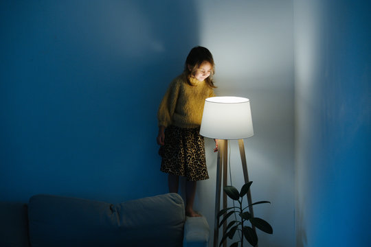 Brunette Little Girl In Yellow Knitted Sweater And Leopard Skirt Standing On A Backseat Of A Couch At Home. She's Looking At Dim Night Light From Above. Her Face Is Lit.