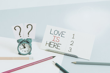 Text sign showing Love Is Here. Business photo text Roanalysistic feeling Lovely emotion Positive Expression Care Joy Mini size alarm clock beside stationary placed tilted on pastel backdrop