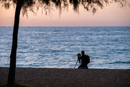 Silhouette of photographer taking photos of sea on the beach during sunset with a tree on the foreground. Barcelona, Spain