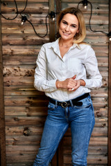 Full-length art portrait of a pretty smiling blonde woman in blue jeans and a white shirt standing in an alternative interior made of wood and lights. The concept of retro, glamor, romance.