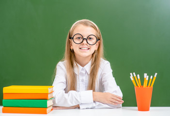 Smiling young girl wearing a eyeglasses sits with books near empty green chalkboard