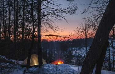 Campfire burns in the snow in the forest hill, on a background of snow covered trees and mountains campfire burning in cold winter. night fire and tent. Hiking Tourism. Flames, background