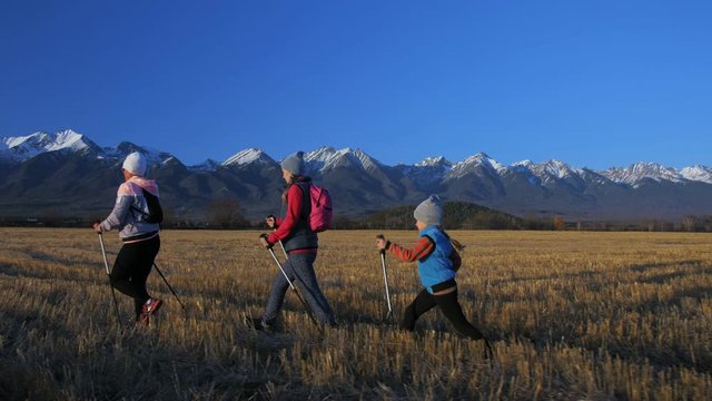 Woman Nordic walking in nature on background of mountains in field. Girls and children use trekking sticks and nordic poles, backpacks. Family travels sports. Kid is learning from mother and