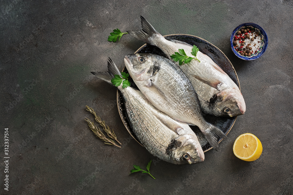 Wall mural fresh raw dorado fish in a plate with lemon, spice and parsley on a dark paint background. top view,