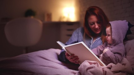 happy mother reading the bedtime stories to infant baby girl in bed at night