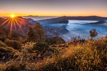 Amazing sunrise over Gunung Bromo volcano in Indonesia. Fog and morning mist in sand valley in Mount Bromo area. Sun rays during beautiful daybreak. Dawn with sunny beams of light and colourful sky.