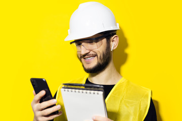 Studio portrait of young smiling man architect, builder engineer, wearing white construction safety helmet, glasses and jacket. Using smartphone and wireless earphones, isolated on yellow background.