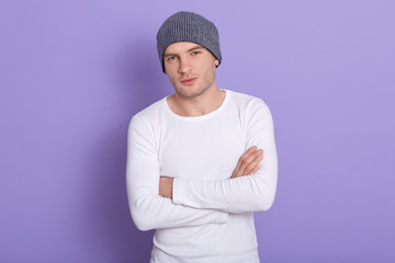 Studio shot of young man standing with hands folded wearing white casual shirt and gray cap. Copy space for advertisment or promotion text. Handsome mle looking directly at camera. People concept.