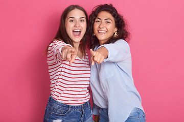 Image of smiling breunette girls posing isolated over pink sudio background, looking amd pointing at camera, female with wavy hair wearing blue shirt, other lady dresses casual shirt with red stripes.