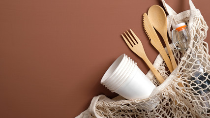 Zero waste concept. Reusable produce bag with wooden cutlery, glass bottle and carton cups on brown background. Eco-friendly, biodegradable, plastic free tableware. Sustainable lifestyle.