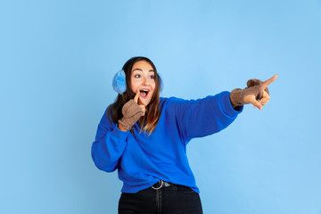 Naklejka premium Pointing. Caucasian woman's portrait on blue studio background. Beautiful female model in warm clothes. Concept of human emotions, facial expression, sales, ad. Winter mood, Christmas time, holidays.