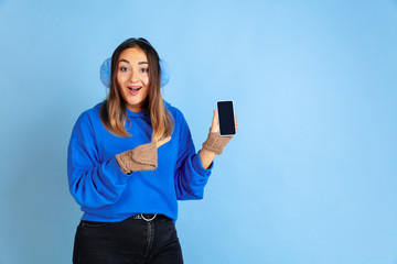 Naklejka premium Showing blank screen. Caucasian woman's portrait on blue studio background. Beautiful female model in warm clothes. Concept of human emotions, facial expression, sales, ad. Winter mood.