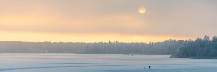 Fantastic winter landscape. Shore of a frozen river covered with snow. The disk of the sun shines through fog and clouds. An orange beautiful dawn dawns upon fishermen in the distance. Web banner.