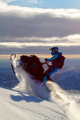 a snowmobile rider jumps in a mountain valley at dawn. sports snow bike with snow splashes and snow trail. bright snowmobile and suit without brands. snowmobilers sports riding. stock photo