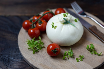 Italian cheese Burrata lies on a round wooden board surrounded by cherry tomatoes and garnished with microgreens. Close up photo on dark wooden table.