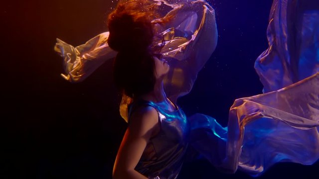 An adult woman in silver clothing underwater swims like a mermaid in the dark under neon lighting