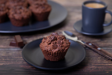 Chocolate muffins on a black plate. Homemade baking. In the background is a cup of coffee and a plate with muffins. Wooden background. Selective focus, close up.