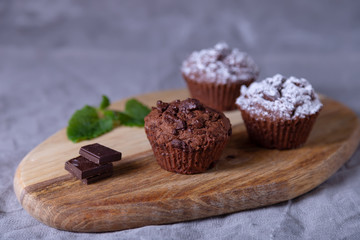 Chocolate muffins on a wooden board, strewed with icing sugar. Homemade baking. In the background is a cup of coffee and a plate with muffins. Wooden background. Selective focus, close up.