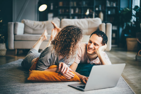 Couple Watching At Computer And Smiling. Two Friends Watching A Video On A Notebook In The Living Room.