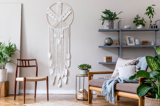 Stylish interior of living room at cozy apartment with brown sofa, coffe table, bookstand, a lot of plants, macrame and elegant accessories. Beige and japandi concept. Modern home staging. Template.