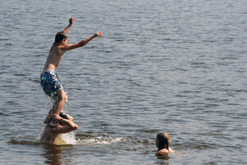 The son jumps into the water from the shoulders of his father. Dad, daughter and son bathe and swim in the lake.
