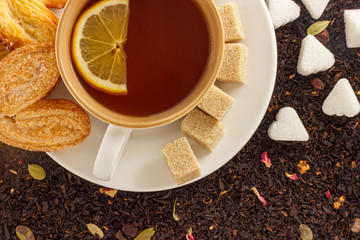 white mug with hot black tea with lemon and cookies on a white saucer on the background of black loose tea top view