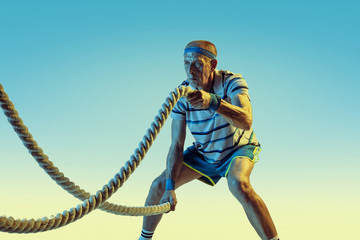 Senior man wearing sportwear training with ropes on gradient background, neon light. Caucasian male model in great shape stays active. Concept of sport, activity, movement, wellbeing, confidence.