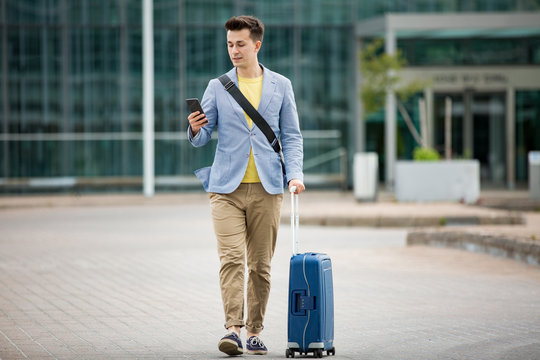 Stylish man standing at airport with smartphone and suitcase, browsing, texting, using mobile app. Business traveling.
