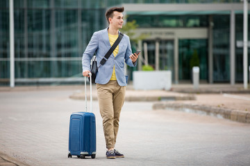 Stylish man standing at airport with smartphone and suitcase, browsing, texting, using mobile app. Business traveling.