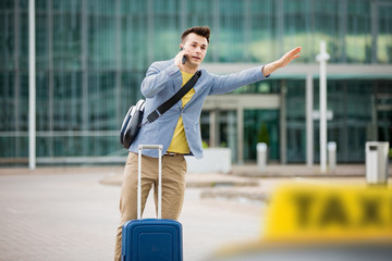 Stylish man standing at airport with suitcase and smartphone, hailing taxi.  Businessman traveling. 