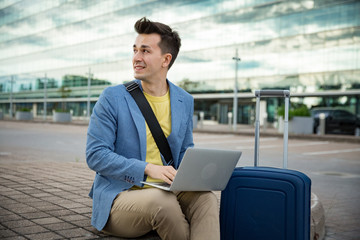 Stylish man sitting at airport with suitcase and laptop, working, typing, browsing. Businessman traveling. 