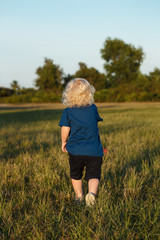 Curly blonde hair little boy toddler child running away in grass outside in his yard chasing ball at sunset golden hour