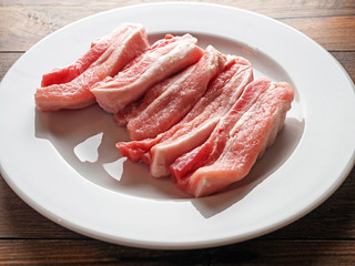 Uncooked raw pork belly slices on a white plate and wooden table.