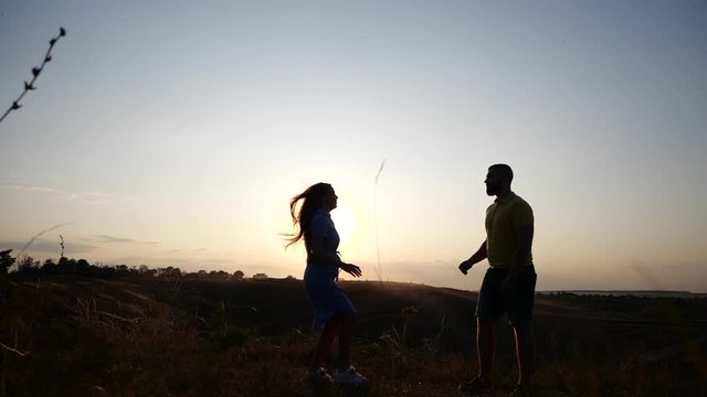 A meeting of a young charming couple in rural landscape at sunset. Silhouette of bearded young man and his girlfriend twirling spinning around at breathtaking golden sunset.