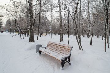 A bench covered with snow in a city Park in winter, with people in the background