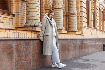 Beautiful young girl in fashionable biege hoodie, elegant gray coat and pants on brick wall background. Walking on the street.