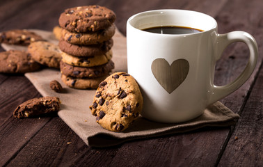 Cup of coffee with heart and chocolate chip cookies on rustic wooden background.