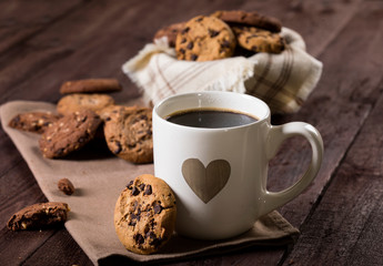 Cup of coffee with heart and chocolate chip cookies on rustic wooden background.