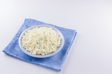 Cottage cheese in a white plate. Fresh cottage cheese on a white background. Homemade curd on a blue napkin in minimalist style. Top view. Copy space