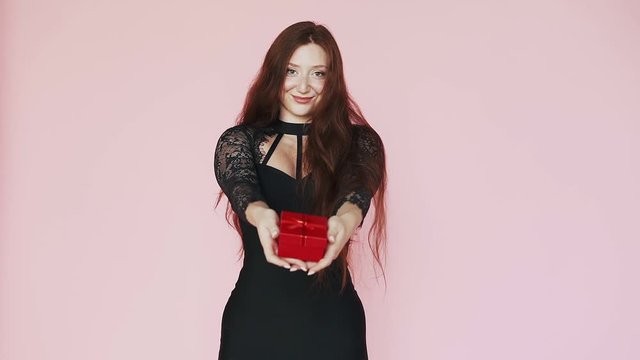 Beautiful red-haired girl in black dress holds out a gift to the camera. Smiles, festive mood. The theme of the 8th of March. Slow motion