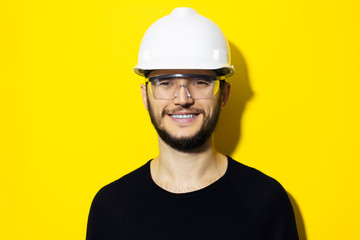 Studio portrait of young smiling man architect, builder engineer, wearing black sweater, white construction safety helmet and glasses isolated on yellow background.