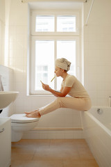 Beautiful young  girl in white towel sitting on a window in the bathroom looking at smartphone and brushes her teeth.